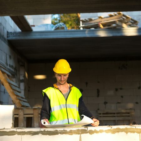 A construction engineer and designer in a yellow hardhat studies a drawing of a building